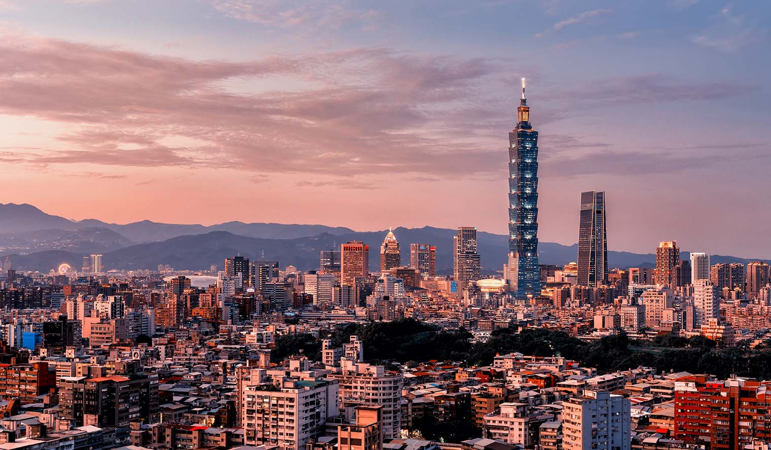 Panoramablick auf die Skyline von Taipeh bei Sonnenuntergang mit dem markanten Taipei 101 Wolkenkratzer, umgeben von urbanen Gebäuden und Bergen im Hintergrund – ein beeindruckendes Motiv für Taiwan Reisen.